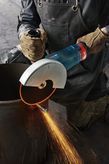 A person wearing safety equipment cuts a metal pipe with a large angle grinder, sparks flying.