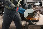 A person wearing safety equipment cuts metal rods with a large circular saw, producing sparks.
