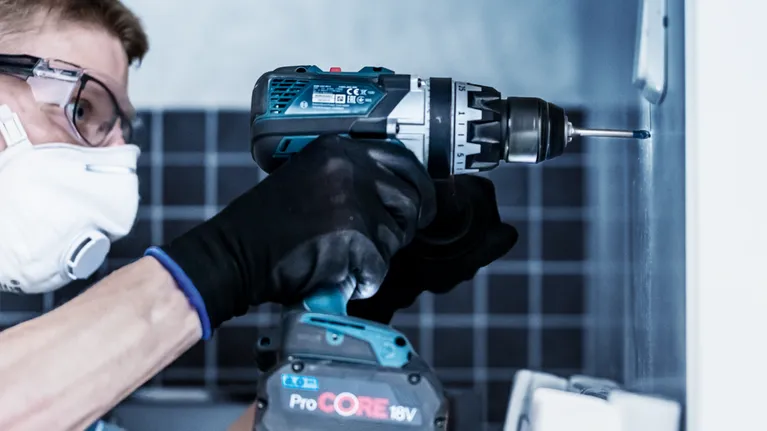 Person wearing safety equipment uses a cordless drill to drive a screw into a wall.
