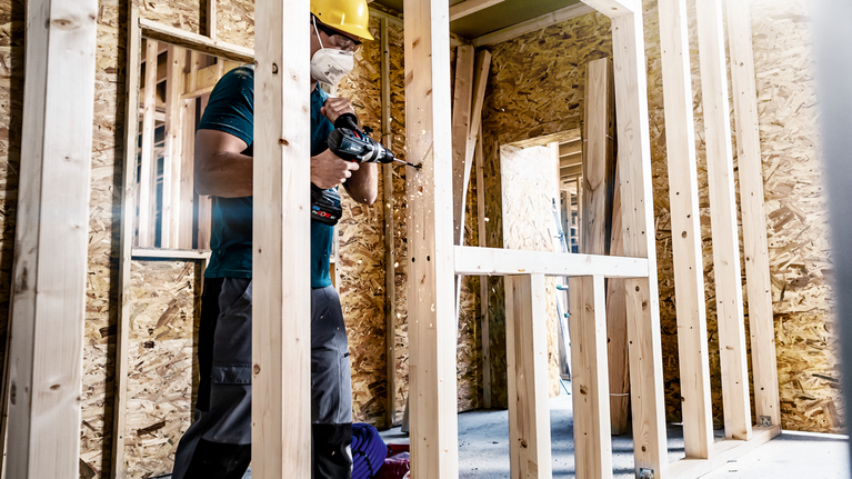 EXPERT Self Cut Speed Spade Drill Bit 40 x 152 mm A person wearing safety equipment drills into a wooden frame at a construction site.