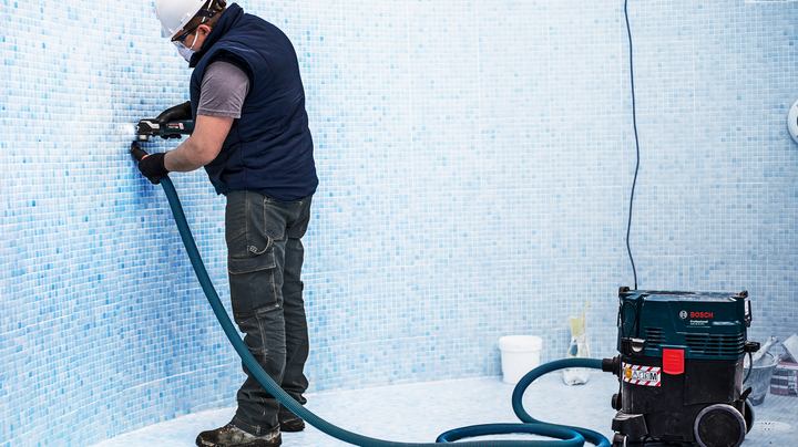 Object #200360601: 2793207_web_001 A worker wearing safety equipment drills into tiled wall, connected to a dust extractor.