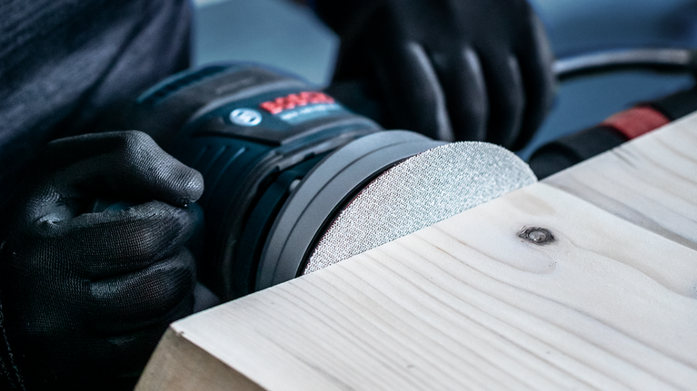 EXPERT M480 Starter Kit for Random Orbital Sanders 125 mm 7-pc Person wearing safety equipment sands a wooden surface with a power tool.