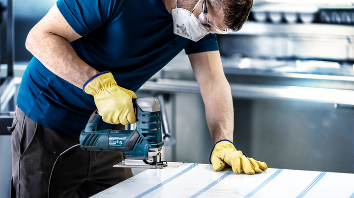 Person wearing safety equipment uses a jigsaw to cut lines in a sheet of material.
