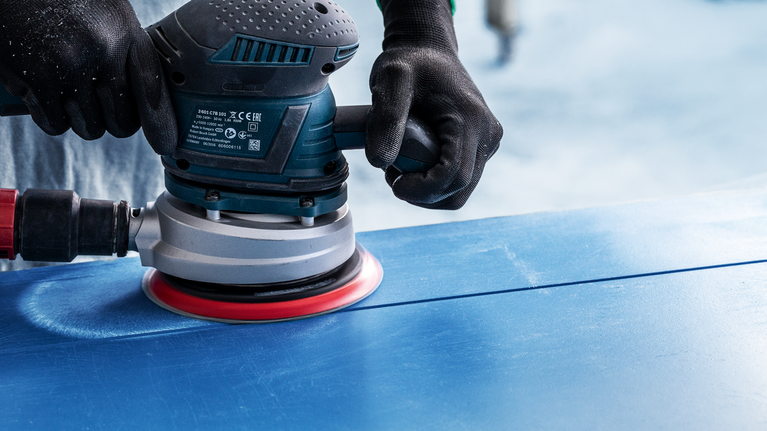 Person wearing safety equipment sanding a blue surface with an electric sander.