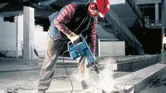 A person wearing safety equipment operates a demolition hammer to break up concrete.