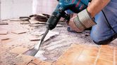A person wearing safety equipment uses a demolition hammer to remove floor tiles.