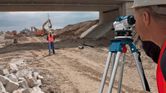 A person wearing safety equipment uses a leveling tool to survey a construction site.