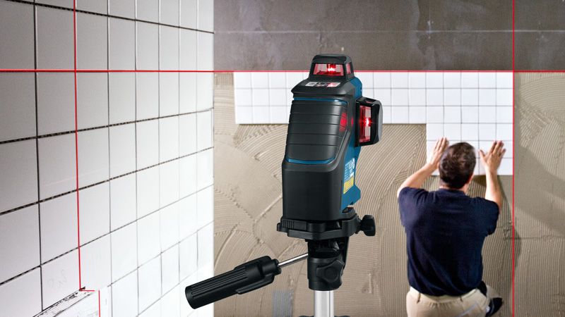 Laser leveling tool projects red lines as a person installs tiles on a wall.