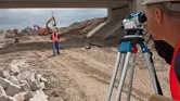 Person wearing safety equipment uses a surveying level tripod on a construction site.