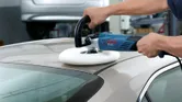 A person polishes a car roof with a power polisher in an indoor garage.