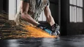 A person wearing safety equipment grinds metal using an angle grinder, sparks flying.