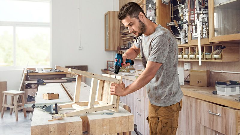 GSR 120-LI A person drills into a wooden frame on a workbench in a woodworking shop.