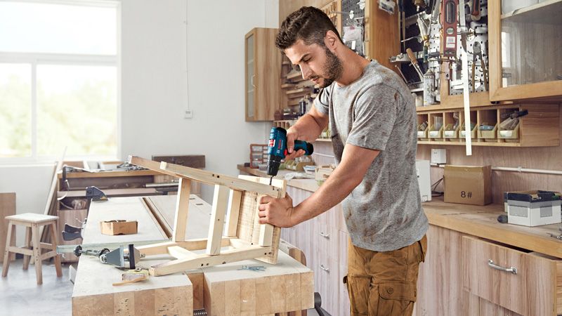A person assembles a wooden chair using a cordless drill in a workshop.