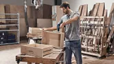 A person wearing safety equipment uses a cordless drill on a wooden drawer in a workshop.