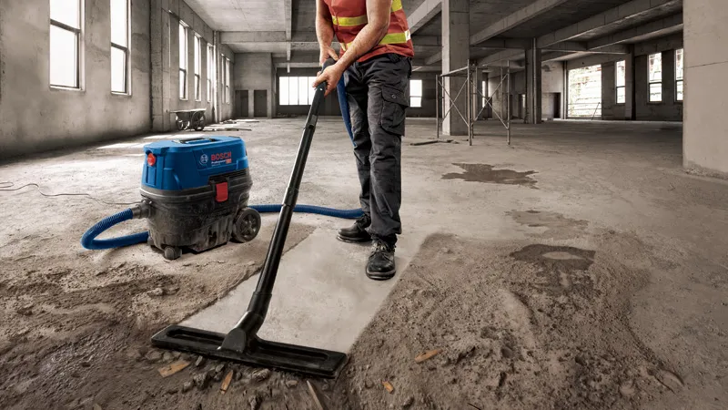 GAS 12-25 PL A person wearing safety equipment vacuums concrete dust in a large unfinished building.