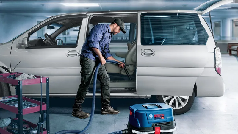 GAS 12-25 PL A person cleans a car interior with a vacuum next to a tool cart in a garage.