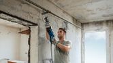 A person wearing safety equipment uses a reciprocating saw to cut a metal pipe indoors.