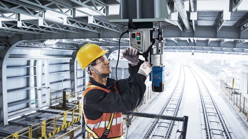 A person wearing safety equipment operates a magnetic drill on a steel beam above train tracks.