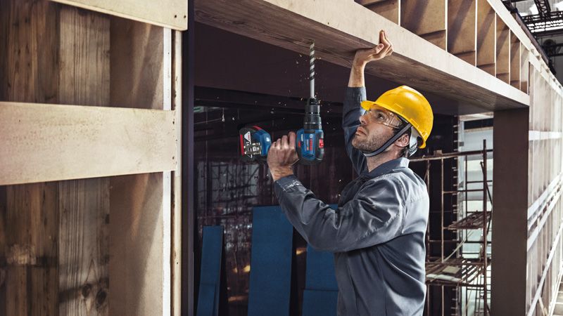 GSB 18V-50 A person wearing safety equipment drills into a wooden beam at a construction site.