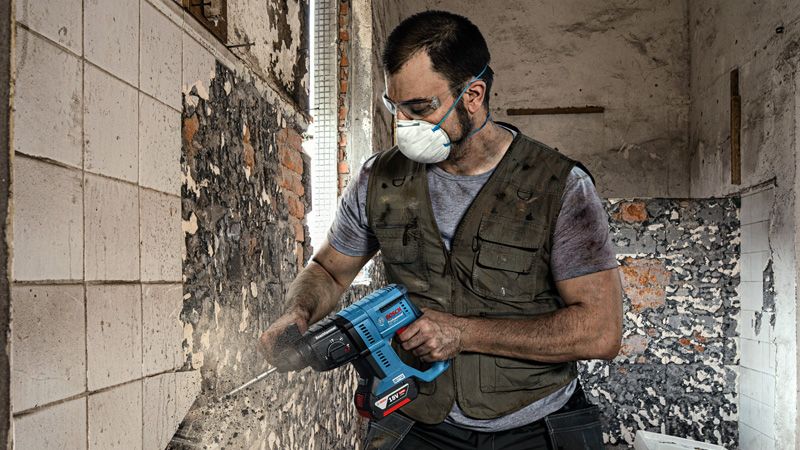 GBH 180-LI A person wearing safety equipment removes tiles from a wall using a power tool.