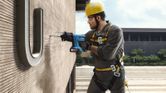 A person wearing safety equipment drills into a building wall while suspended.