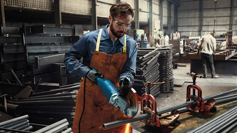 GWS 2200 A person wearing safety equipment cuts metal pipe with a power tool in an industrial workshop.