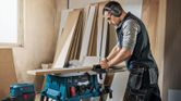 A person wearing safety equipment cuts wood on a table saw in a workshop.