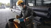 A worker wearing safety equipment grinds a metal pipe with a large angle grinder.