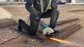 A person wearing safety equipment cuts rebar with an angle grinder, creating sparks.