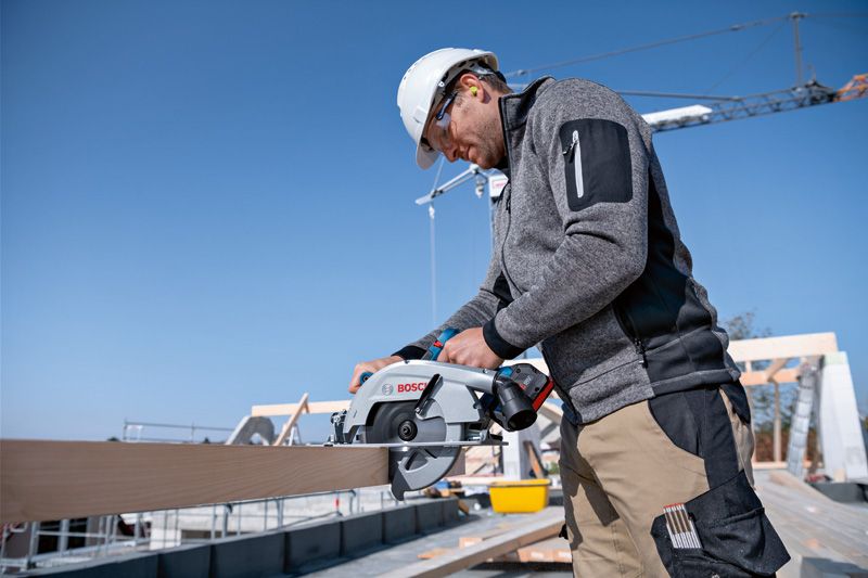 A person wearing safety equipment cuts wood with a cordless circular saw on a construction site.
