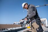 A person wearing safety equipment uses a cordless circular saw to cut a wooden beam.