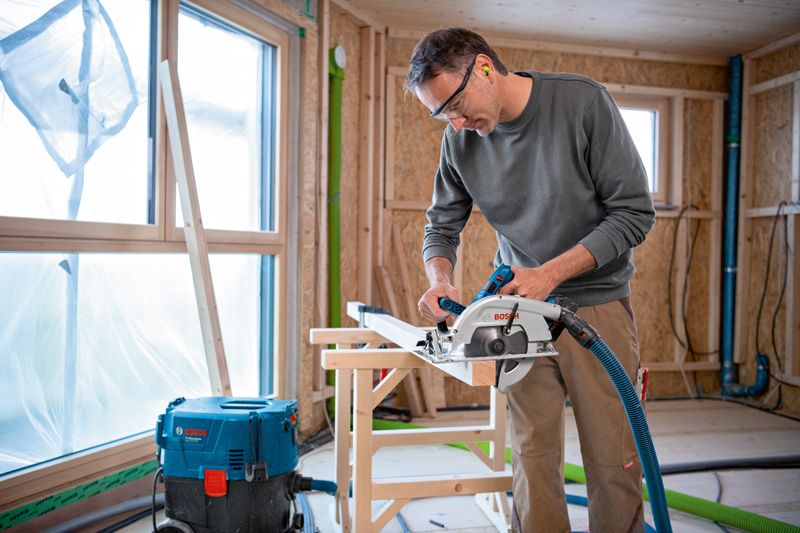 A person wearing safety equipment cuts wood with a cordless circular saw.