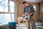 A person wearing safety equipment cuts wood with a cordless circular saw indoors.