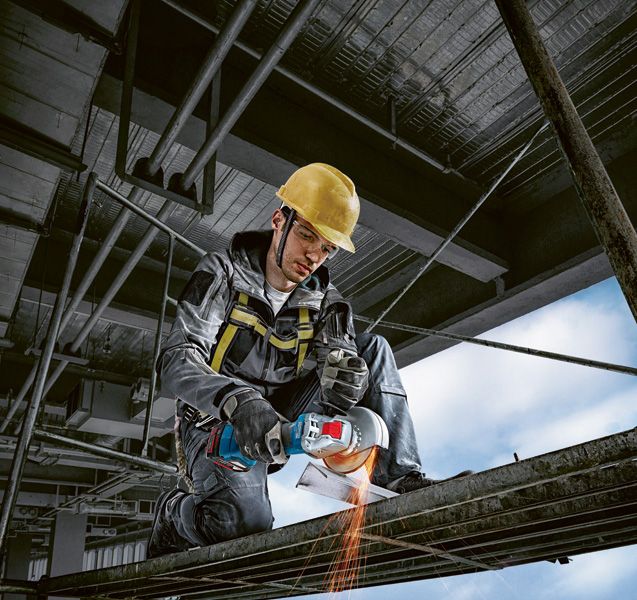 A person wearing safety equipment uses a cordless angle grinder to cut metal.