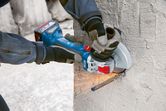 A person wearing safety equipment uses a cordless angle grinder to cut rebar in concrete.