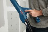 A person adjusts a drywall sander with a hose attached near unfinished drywall.