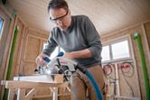 A person wearing safety equipment uses a cordless circular saw to cut wood.