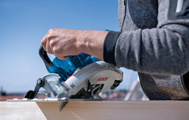 GKS 18V-70 L A person cuts wood outdoors with a cordless circular saw.