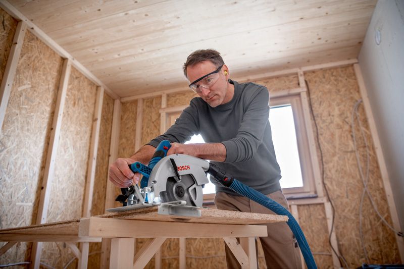 A person wearing safety equipment uses a cordless circular saw to cut wood.
