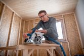 A person wearing safety equipment uses a cordless circular saw to cut wood.