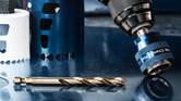 Drill with quick change chuck near a drill bit and hole saws on a reflective surface.