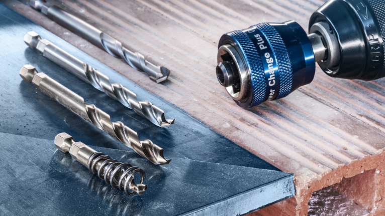 Metal drill bits and a quick-change adapter beside a cordless drill on a brick surface.