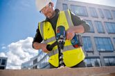 A person wearing safety equipment drills into wood with a cordless drill at a construction site.