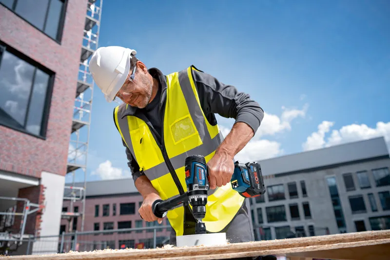 GSR 18V-150 C A person wearing safety equipment drills a hole into wood at a construction site.