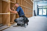 A person wearing safety equipment assembles wooden shelves in a bright room.