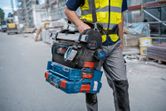 Person wearing safety equipment carries stacked toolboxes and a digital angle measurer at a construction site.