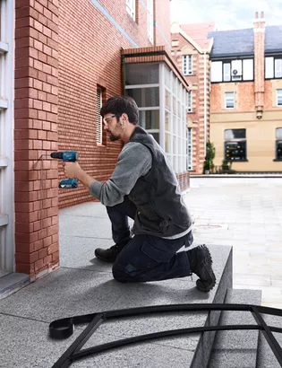Professional Promotion Set of Combo GDX 18V-285 + GSB 185-LI with 2x2.0Ah, GAL 18V-20 in Case A person wearing safety equipment drills into a brick wall on a building exterior.