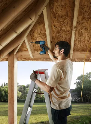 Professional Promotion Set of Combo GDX 18V-285 + GSB 185-LI with 2x2.0Ah, GAL 18V-20 in Case A person on a ladder drills into a wooden roof frame, wearing safety equipment.