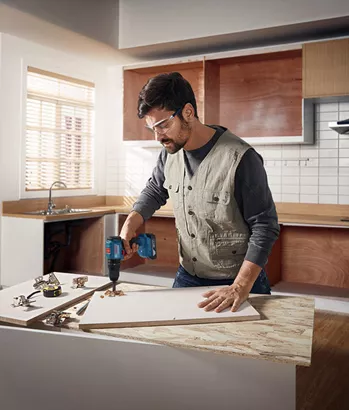 Professional Promotion Set of Combo GDX 18V-285 + GSB 185-LI with 2x2.0Ah, GAL 18V-20 in Case Person wearing safety equipment drills into a wooden board in a kitchen workspace.