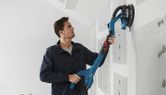 A person sands drywall with a power drywall sander on an unfinished wall.